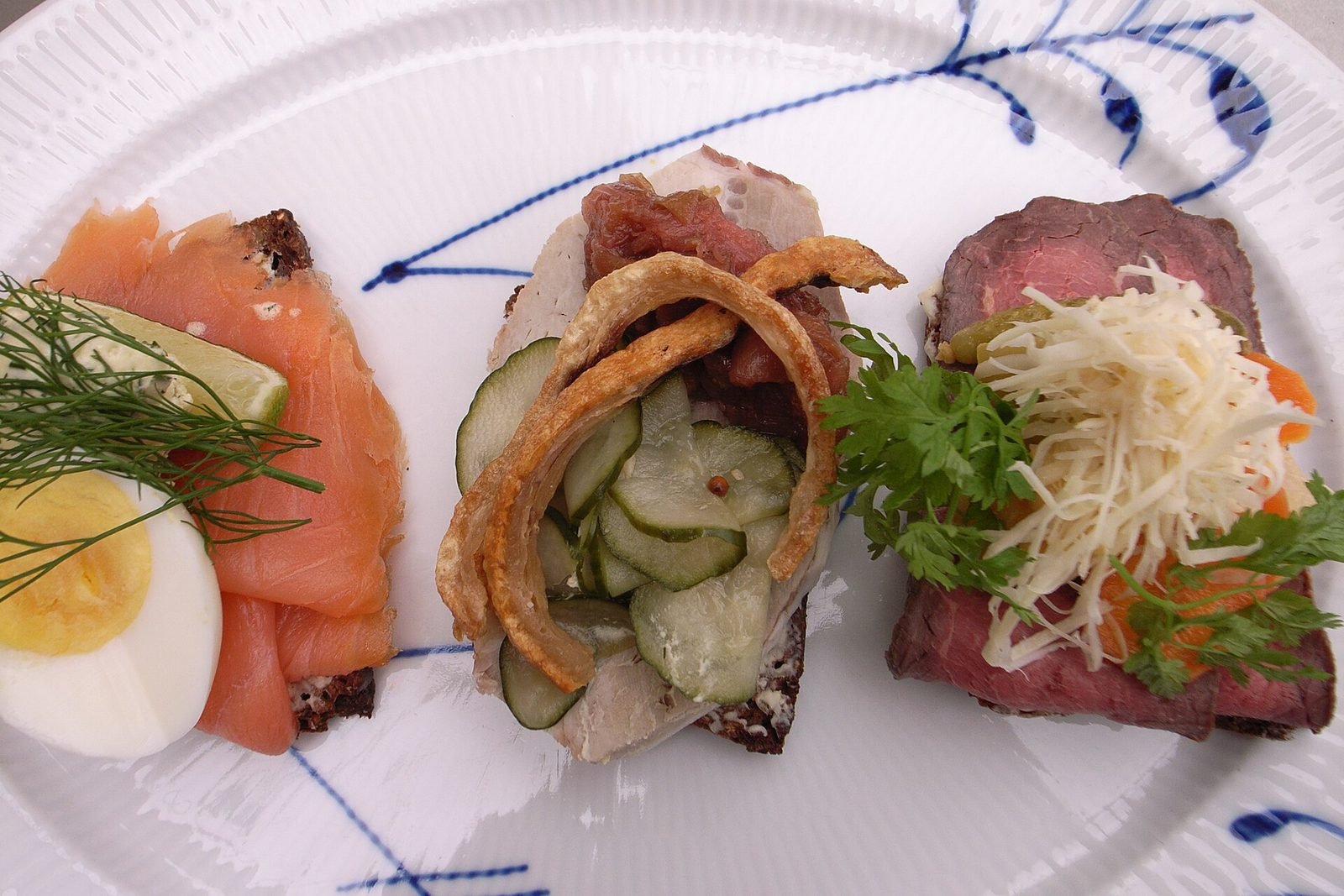 Drei belegte Smørrebrød-Scheiben mit Lachs und Ei, Hering und Roastbeef auf einem blau gemusterten Teller